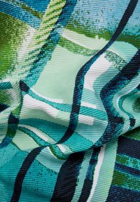 Nahaufnahme eines strukturierten Stoffs mit abstraktem geometrischem Muster in den Farbtönen Blau, Grün, Weiß und Schwarz mit sichtbaren Falten.