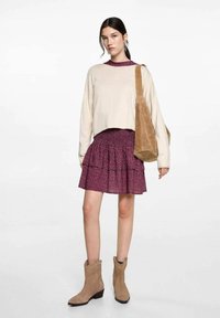 Beiger, verkürzter Pullover, geschichtete lila Blumenrock, beige Wildleder-Stiefeletten und eine braune Schultertasche. Das Outfit zeichnet sich durch weiche Texturen und ein gestuftes Design aus.