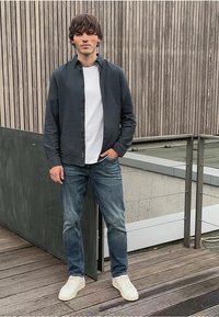 Joven de pie sobre una terraza de madera, vistiendo zapatillas blancas, vaqueros azules, camiseta blanca y una camisa oscura de botones, contra una pared de paneles de madera.