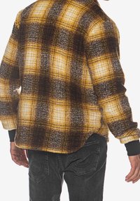 Flannel skjorte med et gult, brunt og gråt ternet mønster, der har en blød struktur, lange ærmer og sorte manchetter.