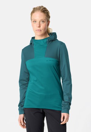 Frau mit blondem Haar, die einen türkisfarbenen Hoodie und schwarze Hose trägt, steht vor einem einfarbigen hellen Hintergrund.