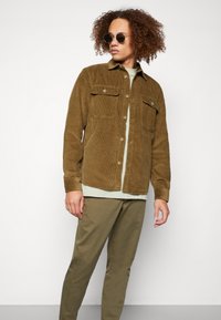 Chaqueta de pana marrón con dos bolsillos en el pecho, combinada con una camisa verde claro y pantalones verde oliva. El modelo lleva gafas de sol redondas.