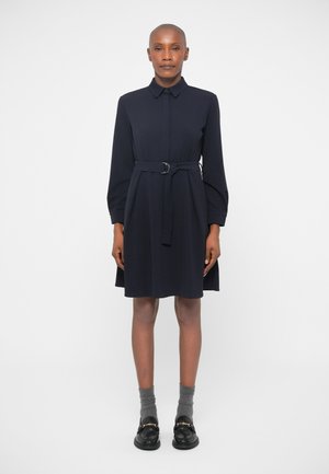 Tamsiai mėlyna suknelė-marškiniai su diržu liemens srityje, ilgomis rankovėmis ir tiesiu siluetu. Nešiojama su juodais batais ir pilkomis kojinėmis. Sklandžiai audinio tekstūra.