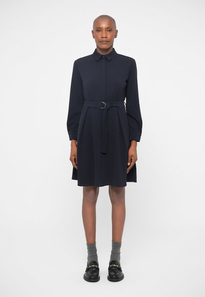 Robe chemise bleu marine avec une taille ceinturée, des manches longues et une coupe droite. Portée avec des chaussures noires et des chaussettes grises. Texture de tissu lisse.