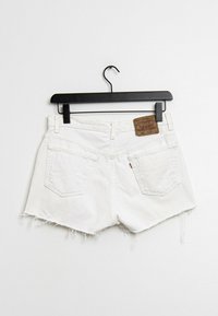 Pantalones cortos de mezclilla blanca con dobladillo deshilachado, dos bolsillos traseros y una etiqueta de marca de cuero marrón en la cintura. Cuelga de un gancho de plástico negro.
