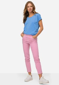 Lichtblauw top met korte mouwen, roze en witte gestreepte broek, witte sneakers, staande met handen in de zakken, neutrale achtergrond.