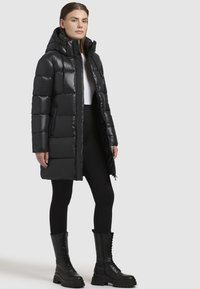 Sort pufferjakke med hætte, med en blank finish, quiltet design, front lynlås og sidelommer. Båret over sorte leggings og støvler.