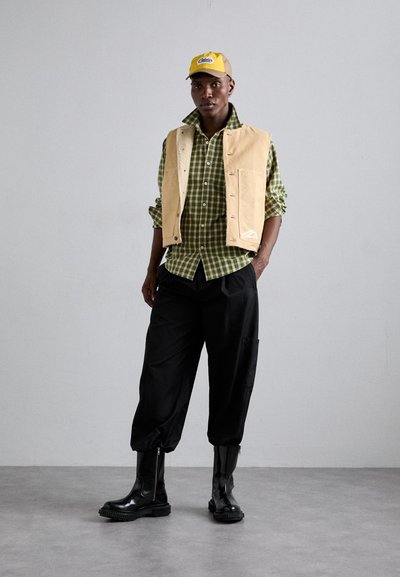 Beige suede vest over a green checkered shirt, black baggy trousers, and shiny black knee-high boots. Yellow cap with logo.