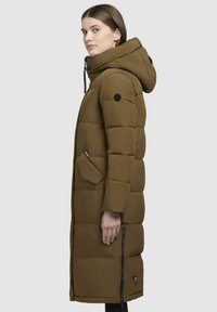 Long olive green puffer coat with a hood, featuring a front zipper, side pockets, and a black logo patch on the sleeve. Textured fabric.