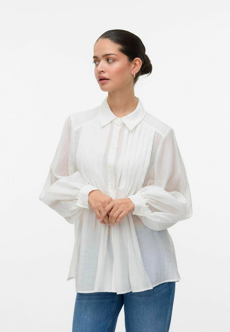 Camicia bianca con bottoni, dotata di un davanti plissettato e un tessuto morbido e trasparente. Presenta un colletto a punta e maniche lunghe a palloncino. Vestibilità casual.