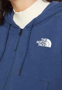 Felpa blue con zip realizzata in morbido tessuto, dotata di cappuccio regolabile con cordino, tasche anteriori e un logo bianco sulla parte sinistra del petto.