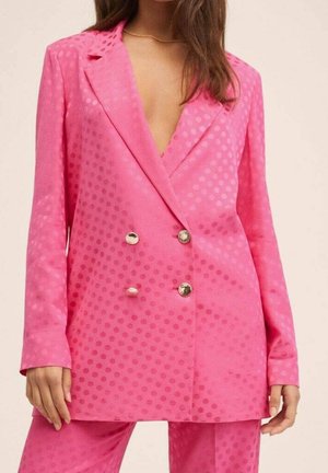 Femme portant un blazer croisé rose vif à pois et un pantalon assorti, mains détendues sur les côtés.
