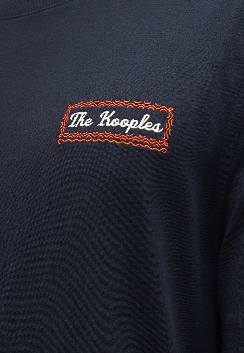 Navy blue cotton t-shirt featuring a rectangular, red embroidered logo with “The Kooples” in white cursive, outlined by a wavy border.