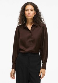 Camicia button-up in satin marrone con colletto, maniche lunghe e dettagli ai polsini. Indossata con pantaloni neri, mostrando una texture e una vestibilità liscia.