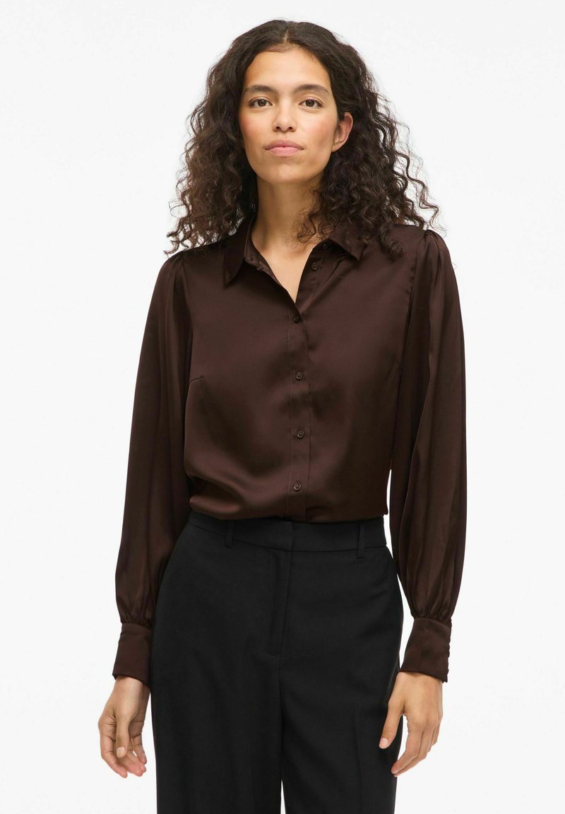 Camicia button-up in satin marrone con colletto, maniche lunghe e dettagli ai polsini. Indossata con pantaloni neri, mostrando una texture e una vestibilità liscia.