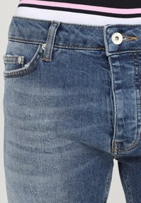 Jeans de mezclilla azul con un diseño clásico. Presentan un bolsillo delantero, cierre con botón metálico y costuras visibles. Textura ligeramente descolorida.