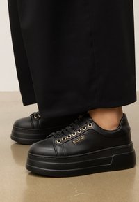 Des baskets plateforme en cuir noir avec des œillets dorés et de la marque, dotées d'une semelle épaisse en noir mat présentant une texture lisse.