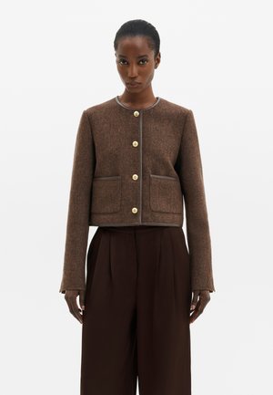 Femme portant une veste courte marron texturée avec des boutons dorés et un pantalon large marron foncé assorti, debout devant un fond uni.
