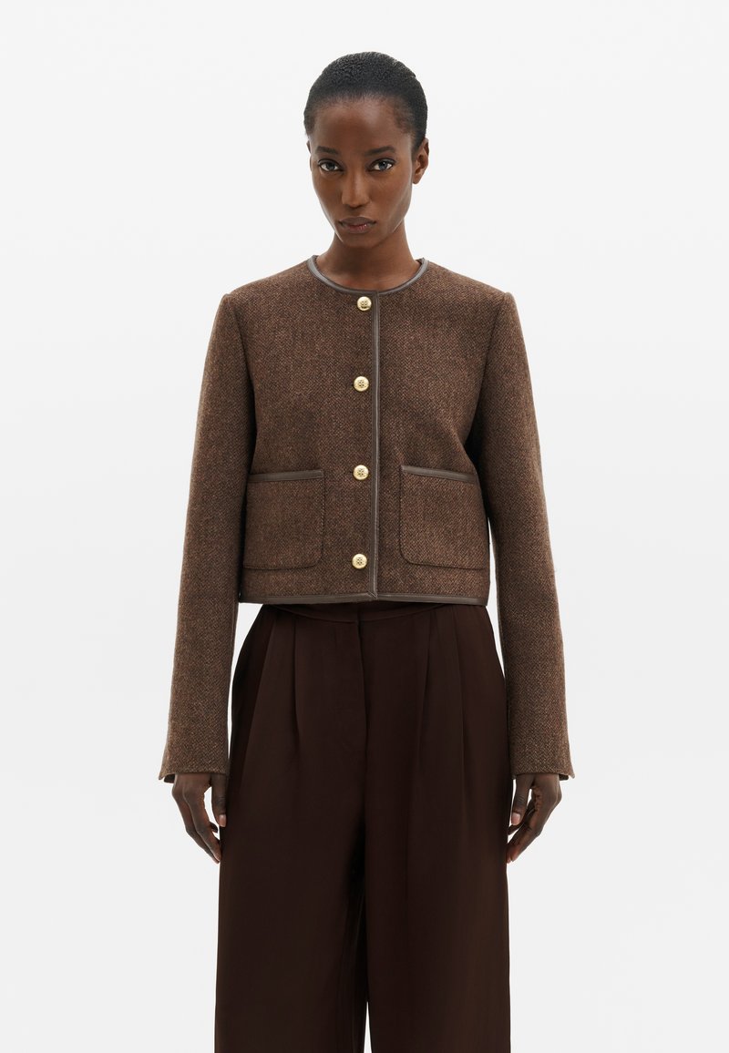 Femme portant une veste courte marron texturée avec des boutons dorés et un pantalon large marron foncé assorti, debout devant un fond uni.