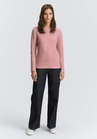 Een vrouw staat en draagt een geribbelde roze longsleeve top, donkere wijde jeans en witte sneakers op een effen lichte achtergrond.