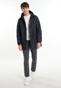 Giacca nera con cappuccio, con chiusura a zip e bottoni, dotata di due tasche frontali. Abbinata a jeans grigi e scarpe da ginnastica bianche.