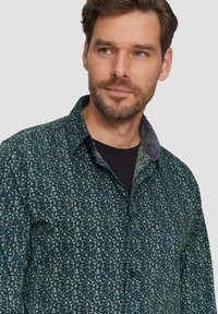 Geprinte shirt met een donkergroene basis en lichtblauwe en zwarte spikkels, met een knoopsluiting bij de kraag en contrasterende details aan de binnenkant van de kraag.