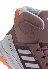 Terrex mid-top athletic shoe in mauve with grey accents. Features a textured fabric upper and a sturdy rubber sole with a vibrant orange tab.