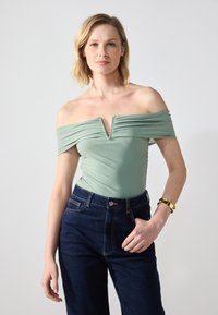 Bodysuit verde menta sin mangas con detalles arrugados y escote en V profundo, combinado con jeans de talle alto de mezclilla oscura. Acento de pulsera dorada.