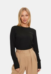 Maglione nero a maniche lunghe con polsini a coste e scollo rotondo, caratterizzato da un logo discreto vicino al petto. Abbinato a pantaloni beige a vita alta.