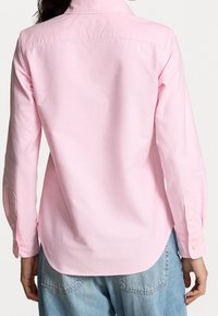 Světle růžová košile s dlouhým rukávem, límečkem, knoflíkovými manžetami a zakřiveným lemem. Viditelné detaily šití a jemná, hladká textura.