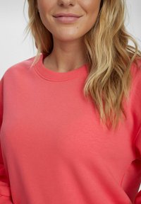 Korallszínű pulóver kerek nyakkal és bordás mandzsettával, puha anyagból készült, sima textúrával és egyszínű kivitelben.