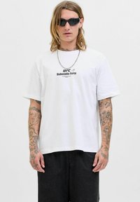 Person med skulderlange hår iført sorte solbriller, en hvid UFC "Undeniable Force" t-shirt, sorte bukser og synlige armtatoveringer.