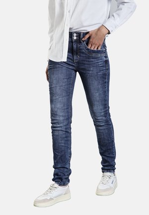 Skinny denim jeans in a dark blue wash, featuring a five-pocket design, slight fading, and silver-tone button hardware, paired with white sneakers.