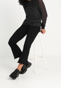 Blouse noire transparente avec de petits motifs blancs, associée à un jean noir évasé et des baskets noires imposantes. Assise sur un tabouret blanc.