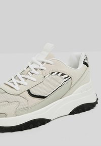 Sport sneakers featuring a light beige mesh upper, black and grey accents, white laces, and a textured, black rubber sole.