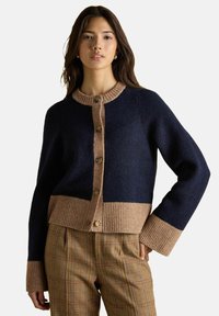 Žena v námorníčko-modrom a béžovom zapínacom kardigáne s rebrovanými manžetami a lemom, v kombinácii s hnedými károvanými nohavicami, postáva na neutrálnom pozadí.