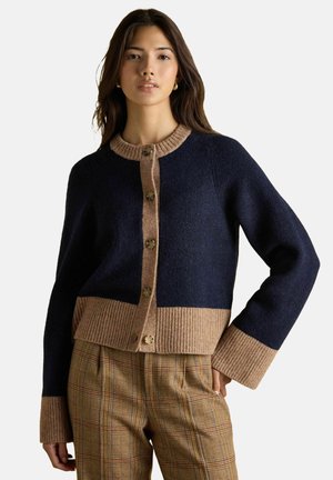Woman wearing a navy and beige buttoned cardigan with ribbed cuffs and hem, paired with high-waisted brown plaid trousers.