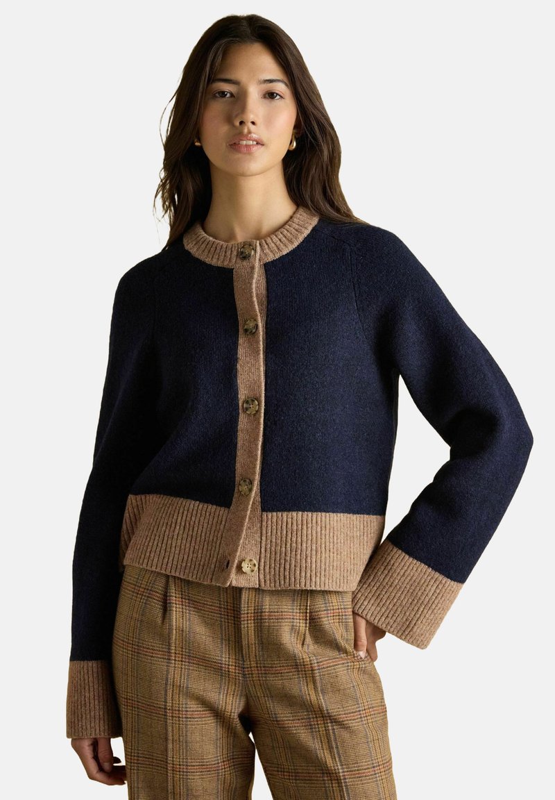 Žena v námorníčko-modrom a béžovom zapínacom kardigáne s rebrovanými manžetami a lemom, v kombinácii s hnedými károvanými nohavicami, postáva na neutrálnom pozadí.