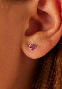 Hartvormige studs in zilver met roze rhinestones, bevestigd aan een close-up van een mensenoor, waarmee de glanzende textuur en het ontwerp worden benadrukt.