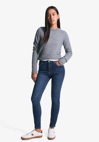 Siv pametana jopica z rebrastim robom, v kombinaciji s temno modrimi skinny kavbojkami. Bele superge s rjavimi poudarki. Model stoji z rokama v žepih.