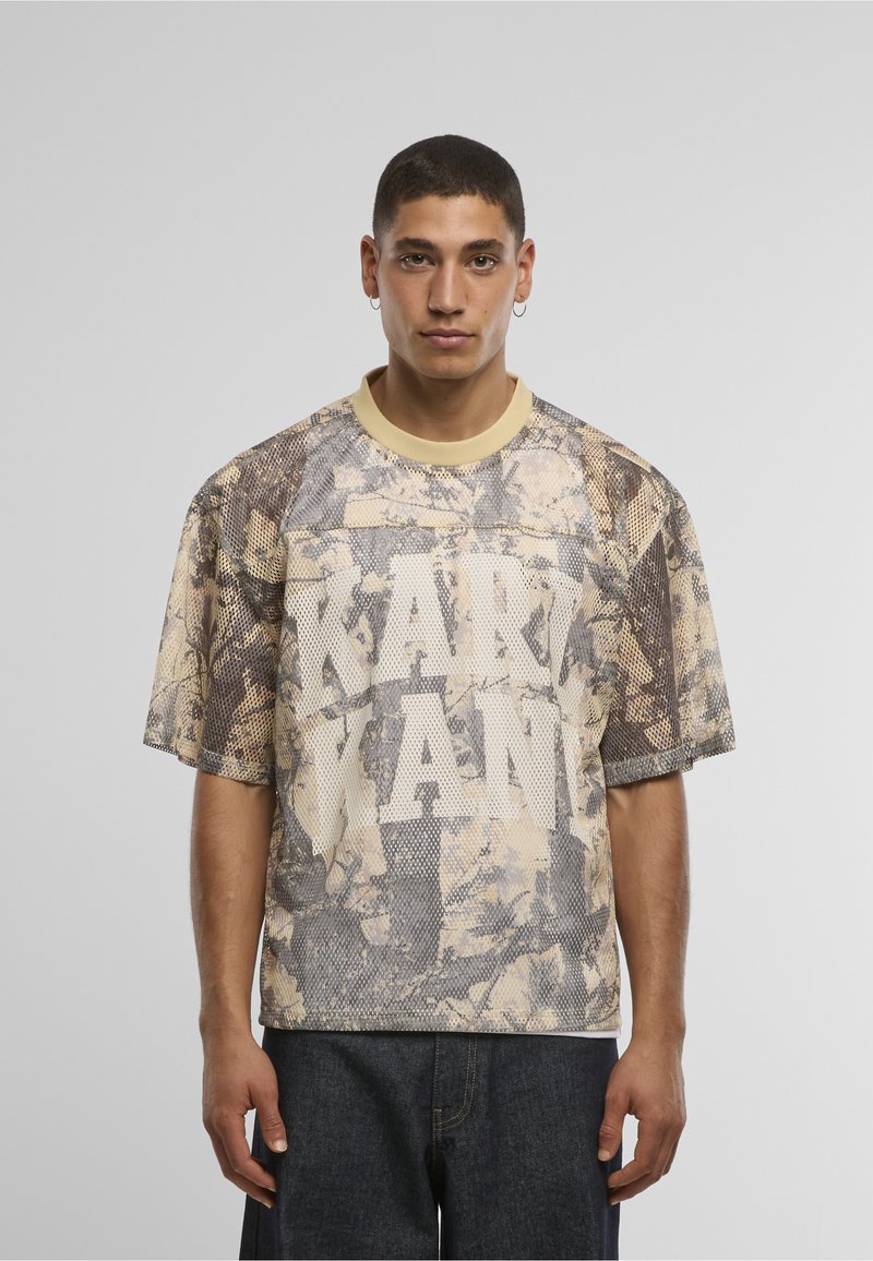 Beige ja ruskea verkkopaita, jossa on maastokuvio. Pyöreä kaulus ja edessä näyttävät valkoiset kirjaimet "KANYE". Lyhyet hihat.