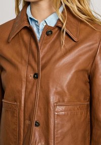 Giubbotto in pelle marrone caratterizzato da un colletto classico, chiusura frontale con bottoni e due tasche inferiori. Texture liscia con una leggera lucentezza.