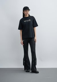 Γυναίκα που φοράει μαύρο καπέλο, oversized μαύρο t-shirt «Off-White», μαύρο cargo παντελόνι με αγκράφες και φερμουάρ, και μαύρα παπούτσια, στέκεται μπροστά σε απλό φόντο.