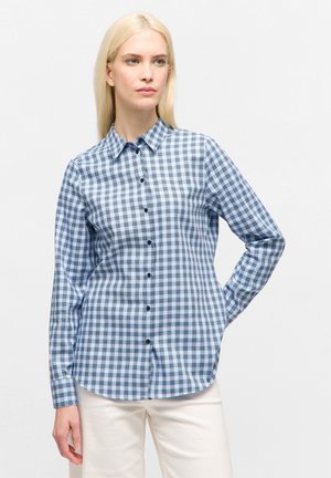 Donna con lunghi capelli biondi che indossa una camicia a quadri blu e bianca con bottoni e pantaloni bianchi, in piedi con una mano dietro la schiena.