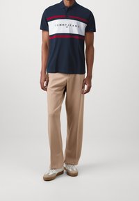 Camisa polo azul marino con rayas horizontales blancas y rojas, que presenta el logo "TOMMY JEANS". Combinada con pantalones beige de corte holgado y zapatillas informales.