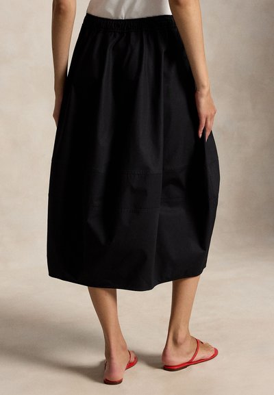 Γυναίκα που φορά μαύρη midi φούστα με ελαστική μέση, συνδυασμένη με κόκκινα σανδάλια με επίπεδη σόλα, στέκεται μπροστά σε ουδέτερο φόντο.