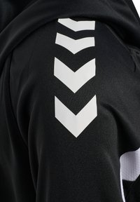 Tissu noir avec une rangée verticale de cinq flèches chevrons blanches pointant vers le bas le long d'une manche ou d'une jambe de pantalon.