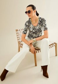 Vrouw in een gebloemd topje, witte broek, bruine haklaarzen, gouden sieraden en een zonnebril, zittend op een houten stoel met een geweven zitting.