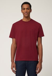 T-shirt in cotone borgogna con collo a giro e maniche corte, con un piccolo logo nero sul lato sinistro del petto, abbinato a jeans in denim scuro.