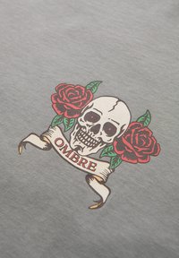 Crâne aux yeux rouges creux centré entre deux roses rouges et des feuilles vertes, au-dessus d'une bannière enroulée portant l'inscription "OMBRE" sur un tissu gris.